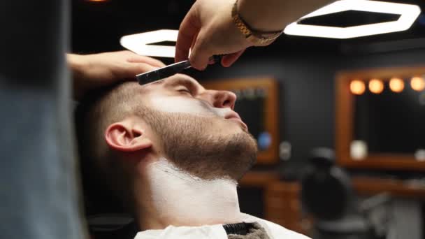 Profesionální holič holí vousy zákazníků břitvou. Vousy ostříhané staromódní čepelí v holičství. Pohledný chlapík, co se holí ve studiu. Detailní záběr. — Stock video
