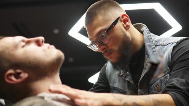 Professzionális borbély borotvával borotválja az ügyfelek szakállát. Szakállas vágás régimódi pengével a fodrászatban. Jóképű macsó férfi borotválkozik a stúdióban. Közelkép.. — Stock videók