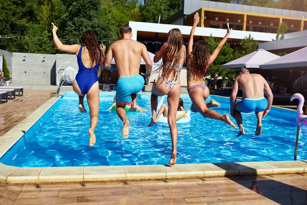 Boldog barátok ugrás és fröccsenő medence felfújható úszók luxus üdülőhely. A fürdőruhás fiatalok ünnepelnek a magánnyaralóban a napsütéses napon. A dögös lányok vízbe ugranak.. — Stock Fotó