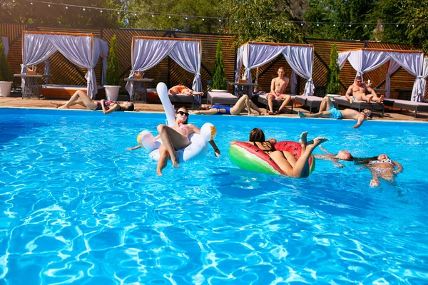 Grupo de amigos que se relajan en la piscina privada de la villa mienten en el sol en el flamenco inflable, el cisne, el colchón. Los jóvenes se relajan con pisos en un resort de lujo en un día soleado. Bikini chicas tomando el sol. —  Fotos de Stock