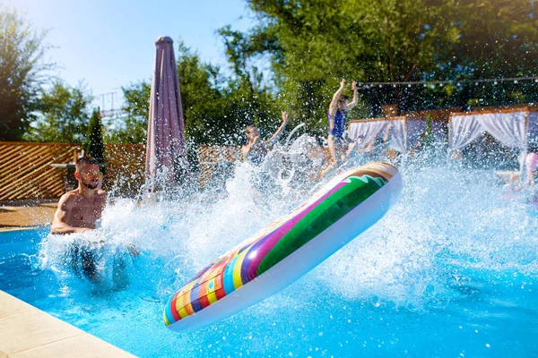 Amigos felizes pulando e salpicando na piscina com flutuadores infláveis em resort de luxo. Jovens em roupa de banho fazendo festa em casa de férias privada no dia ensolarado. Meninas quentes pular na água. — Fotografia de Stock