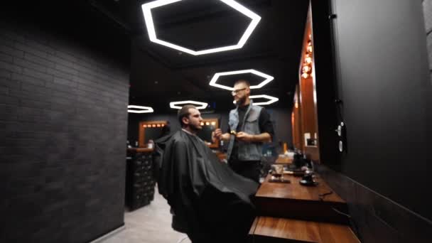 Trendy barber cuts bearded mans hair with a clipper in barbershop. Mens hairstyling and hair cutting in salon. Grooming the hair with trimmer. Hairdresser doing haircut in retro hair salon. Dolly in — Stock Video
