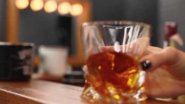 Mujer experimentada sommelier agitando un vaso con whisky y inhalando aroma a alcohol. La mano femenina mueve el vidrio con brandy caro en el mostrador de la barra de madera en la barbería . — Vídeos de Stock