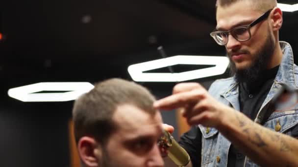 Peluquero en gafas corta el pelo de los hombres barbudos con un cortador en la barbería. Hipster peinado y corte de pelo en el salón. Aseo el cabello con trimmer. Peluquería haciendo corte de pelo en peluquería retro . — Vídeos de Stock