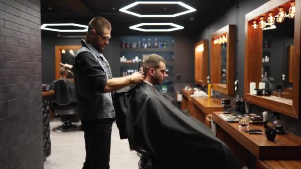 El peluquero moderno corta el cabello de los hombres barbudos con un cortador en la barbería. Para hombre peinado y corte de pelo en el salón. Aseo el cabello con trimmer. Peluquería haciendo corte de pelo en peluquería retro. Seguimiento — Vídeo de stock