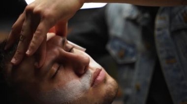 Profesyonel berber, müşteri sakalını ustura ile tıraş eder. Berber dükkanında eski moda bıçakla kesilmiş bir sakal. Yakışıklı maço adam stüdyoda sakalını kestiriyor. Yakın çekim..