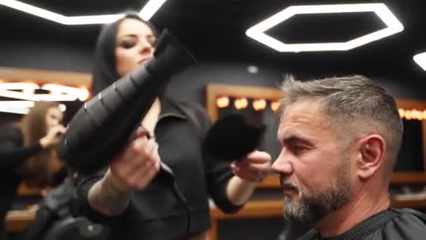 A fodrász nő hajszárítóval és hajkefével dolgozik a fodrászatban. A tetovált borbély lány férfi hajat szárít vágás után. Ügyfél szolgálja fel a vintage haj stúdió. — Stock videók