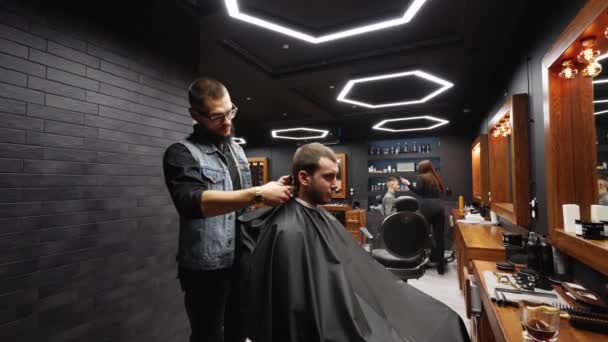 Barbeiro moderno corta barbudo homem cabelo com um cortador de cabelo na barbearia. Cabeleireiro masculino e corte de cabelo no salão. Arrumando o cabelo com aparador. Cabeleireiro fazendo corte de cabelo no salão de cabeleireiro retro. Rastreamento — Vídeo de Stock