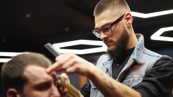 Peluquero en gafas corta el pelo de los hombres barbudos con un cortador en la barbería. Hipster peinado y corte de pelo en el salón. Aseo el cabello con trimmer. Peluquería haciendo corte de pelo en peluquería retro . — Vídeos de Stock