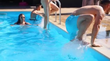 Arkadaşlar yüzme havuzundan sürünerek çıkarlar. Islak mayo giyen mutlu gençler sıcak güneşli bir günde tatil köyündeki partiden sonra havuzdan çıkıyorlar. Kız merdivenden tırabzanlarla çıkıyor. Yavaş çekim.