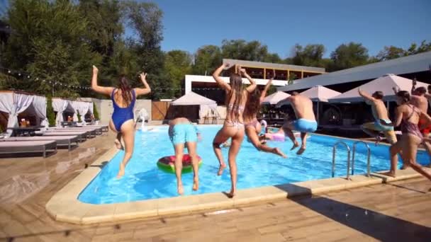 Szczęśliwi przyjaciele skaczą i pluskają się w basenie z nadmuchiwanymi pływakami w luksusowym kurorcie. Młodzi ludzie w strojach kąpielowych urządzający imprezę w prywatnej willi wakacyjnej. Gorące dziewczyny wskakują do wody. Zwolniony ruch. — Wideo stockowe