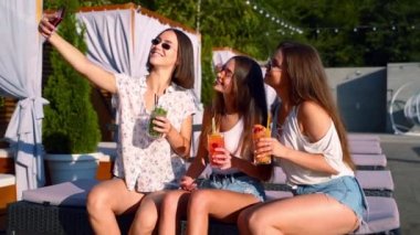 Güneşli bir günde havuz partisinde güneşli bir yerde oturmuş renkli kokteyllerle akıllı telefonuyla selfie çeken gülümseyen kızlar. Lüks villada arkadaşlarıyla tropikal tatilde fotoğraf çeken bir kadın. Yavaş çekim.