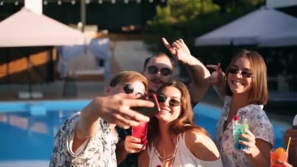 Amigos sorridentes tirando selfie com smartphone na festa da piscina com coquetéis coloridos frescos à beira da piscina no dia ensolarado de verão. Mulher tirando foto na villa de luxo em férias tropicais . — Vídeo de Stock