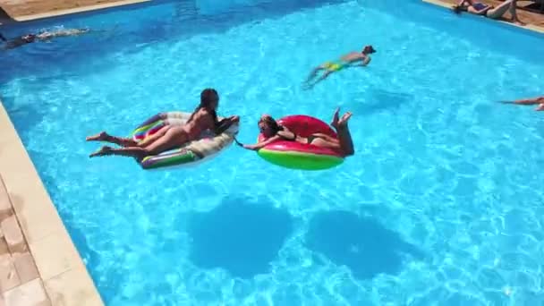 Flygfoto av vänner som har fest i poolen med uppblåsbar flamingo, svan, madrass. Glada ungdomar viftar med händerna mot drönarkameran på lyxanläggningen. Utsikt uppifrån. Flickor i bikini som solar. — Stockvideo