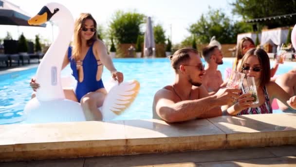 Друзья с коктейлями в бассейне виллы. Счастливые молодые люди в купальниках танцуют, клубятся с надувным фламинго, лебедем, матрасом на роскошном курорте в солнечный день. Медленное движение — стоковое видео