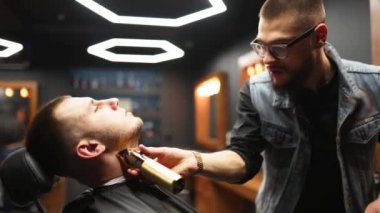 Gözlüklü berber berber dükkanında tıraş makinesiyle adamın sakalını kesiyor. Kuaförde saç ve saç kesimi. Kırpıcı ile saçı tımar etmek. Hippi kuaför retro kuaförde saç kesimi yapıyor..