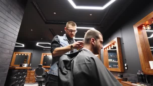 Trendy barber cuts bearded mans hair with a clipper in barbershop. Mens hairstyling and hair cutting in salon. Grooming the hair with trimmer. Hairdresser doing haircut in retro hair salon. Dolly — Stock Video