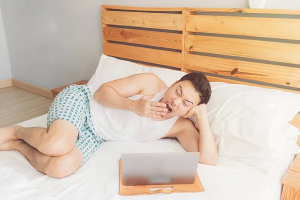 Sleepy hombre está trabajando con su portátil en su acogedora cama. Concepto de aburrido estilo de vida freelancer . — Foto de Stock