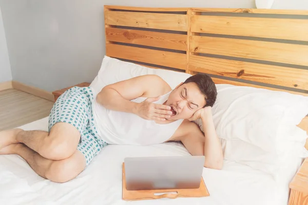 졸린 남자는 그의 아늑한 침대에 자신의 노트북과 함께 일하고 있다. 지루한 프리랜서 라이프 스타일의 개념. — 스톡 사진