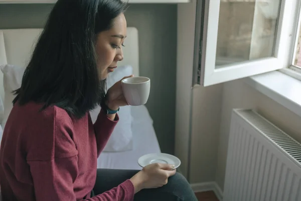 Femme boit du café chaud et regarde par la fenêtre dans son appartement . — Photo