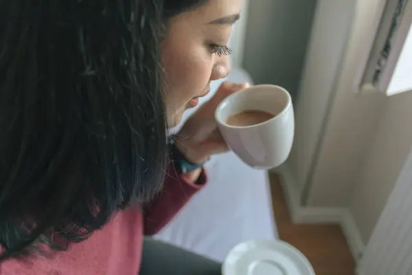 Asian woman is drinking hot coffee and looking out to the window in her apartment.