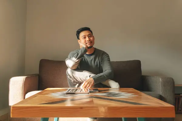 Asian man sits on the sofa in his living room, feeling relax in the morning.