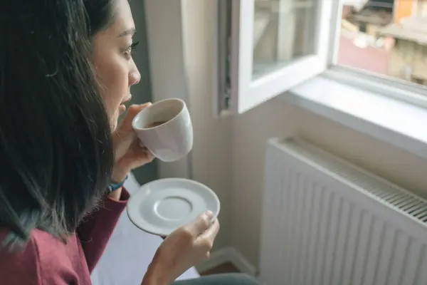 Asian woman is drinking hot coffee and looking out to the window in her apartment.