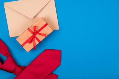 Composition of red neck tie, gift box with bow and brown craft paper envelope on blue cyan background.