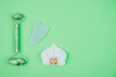 Green face roller and gua sha massager made from natural jade nephritis stone with orchid flower. Facial massage concept. clipart
