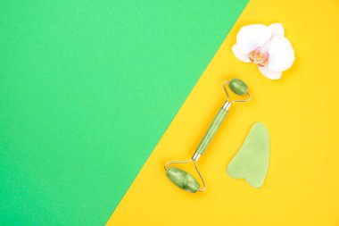 Top view of green face roller and gua sha massager made from natural jade nephritis stone with orchid flower over green and yellow background. Facial massage concept. clipart