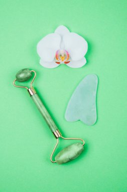 Green face roller and gua sha massager made from natural jade nephritis stone with orchid flower. clipart