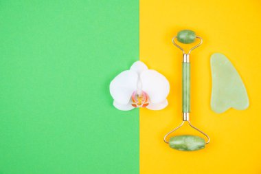 Top view of green face roller and gua sha massager made from natural jade nephritis stone with orchid flower over green and yellow background. Facial massage concept. clipart