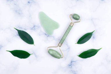 Green face roller and gua sha massager made from natural jade nephritis stone with green leaves over marble background. clipart