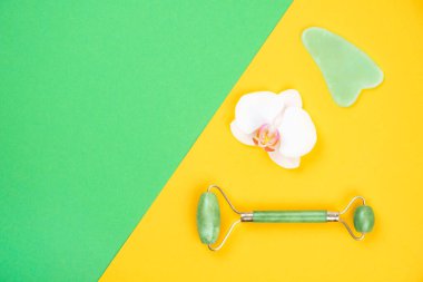 Face roller and gua sha massager natural jade nephritis stone with orchid flower over green and yellow background. clipart