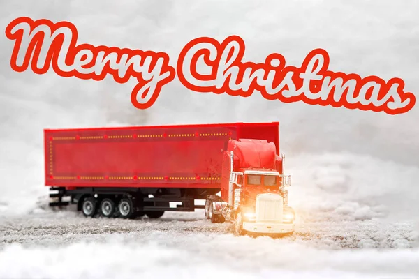 Lettering Merry Christmas. A red festive toy truck with a trailer shines headlights ahead. The tractor rides in the snow. Christmas holidays are coming. Front side view