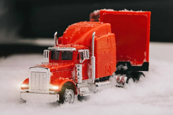 A red celebratory toy truck with a trailer shines headlights ahead. The tractor rides in the snow. Christmas holidays are coming. Front view