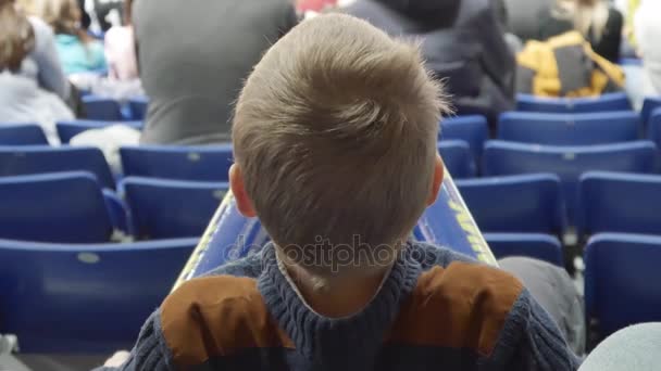 Хлопчик сидить на стадіоні і дивиться спорт - футбол або баскетбол — стокове відео