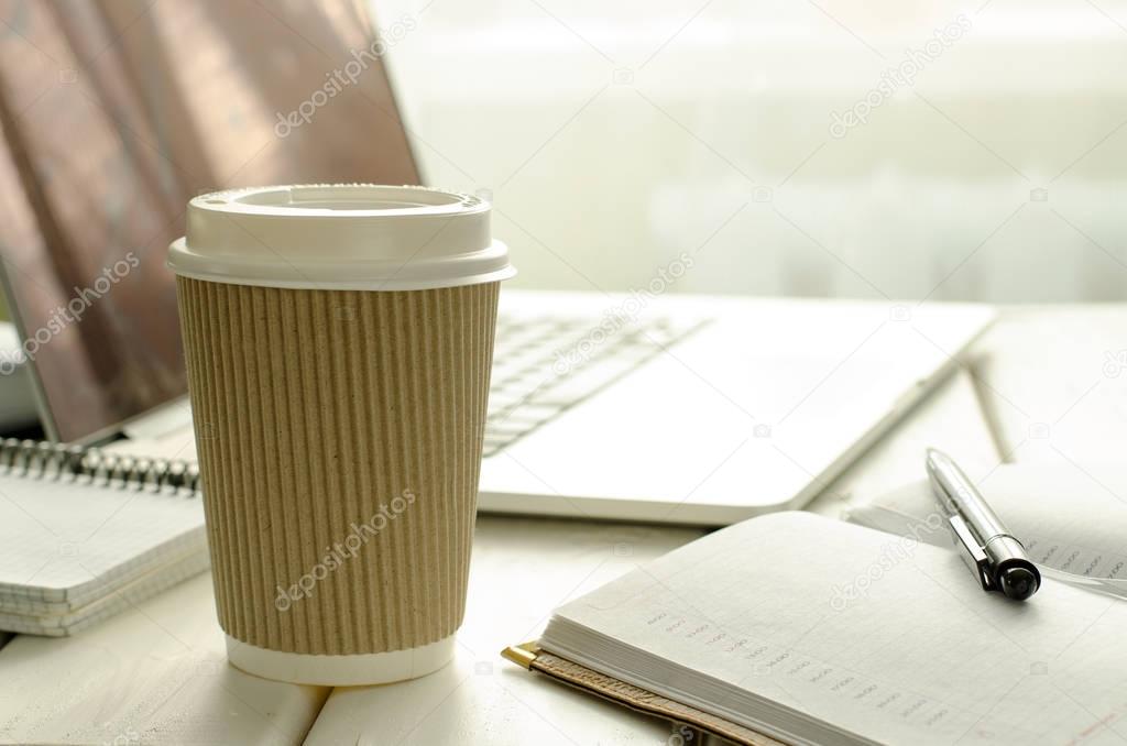 Paper cup of coffee on work-table with office stuff, notepad, laptop, pen and coffee cup