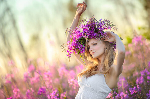 Young beautiful blonde girl with a flower wreath on her head gathers pink flowers in the spring blooming field at sunset.