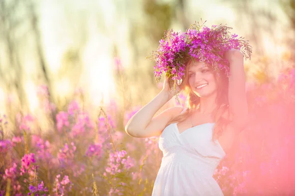 若いです美しいですブロンド女の子とともに花花輪で彼女の頭の上に集まりますピンクの花で春の開花フィールドで日没 — ストック写真