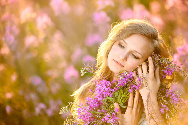 Young beautiful blonde girl gathers pink flowers in the spring blooming field at sunset. The concept of beauty, freshness, youth and health for advertising natural cosmetics.