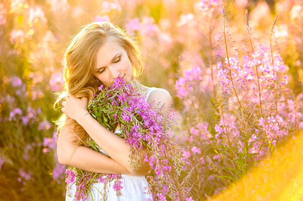 Young beautiful blonde girl gathers pink flowers in the spring blooming field at sunset. The concept of beauty, freshness, youth and health for advertising natural cosmetics.