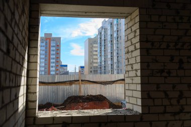 Ev yapımı. Tamamlanmamış bir evin penceresinden çok katlı yeni binaların ve mavi gökyüzünün manzarası. İnşaat sektöründe kriz.
