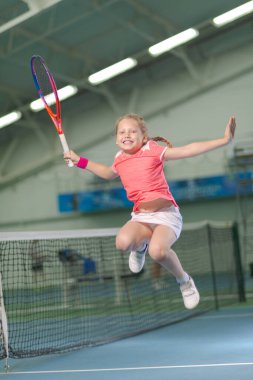 Kapalı tenis kortundaki küçük bir kız tenis maçını kazandığı için mutlu. Spor kulübünde tenis raketi ve topu olan küçük bir kız. Küçük çocuk için antrenman. Çocuk çalmayı öğreniyor.