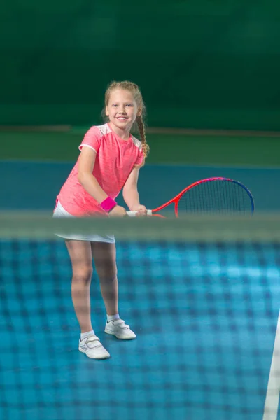 A girl plays tennis on an indoor tennis court. Little girl with tennis racket and ball in sport club. Active exercise for kids. Training for young kid. Child learning to play.