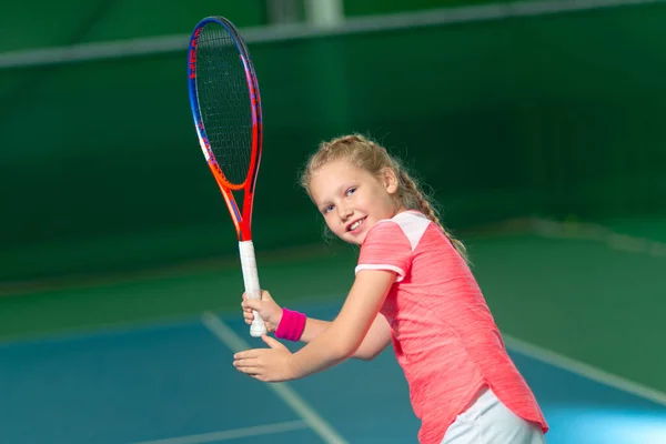 A girl plays tennis on an indoor tennis court. Little girl with tennis racket and ball in sport club. Active exercise for kids. Training for young kid. Child learning to play.