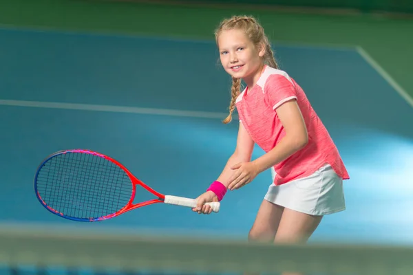 Bir kız kapalı tenis kortunda tenis oynuyor. Spor kulübünde tenis raketi ve topu olan küçük bir kız. Çocuklar için aktif egzersiz. Küçük çocuk için antrenman. Çocuk çalmayı öğreniyor.