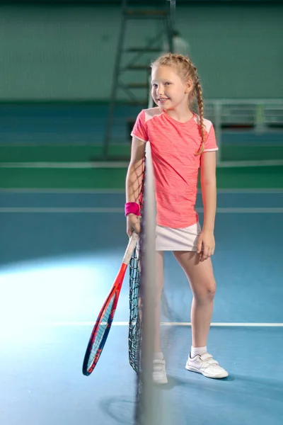 A girl plays tennis on an indoor tennis court. Little girl with tennis racket and ball in sport club. Active exercise for kids. Training for young kid. Child learning to play.