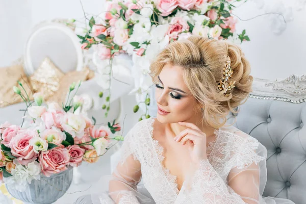 Hermosa chica con el pelo rubio en un peignoir elegantemente se sienta en el sofá rodeado de flores —  Fotos de Stock