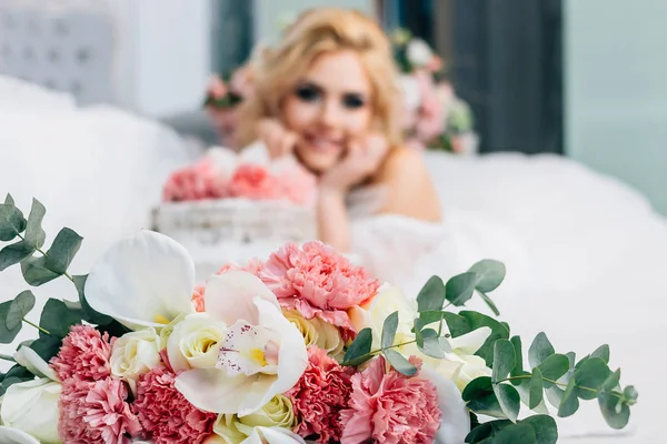 A wedding bouquet in the foreground lies on the floor in a bright room and a beautiful bride with a wedding cake in the background